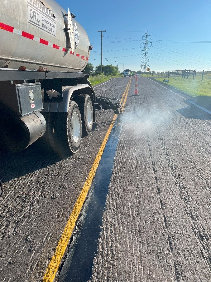 A large truck sprays a thing band of liquid asphalt on the right of a road centerline