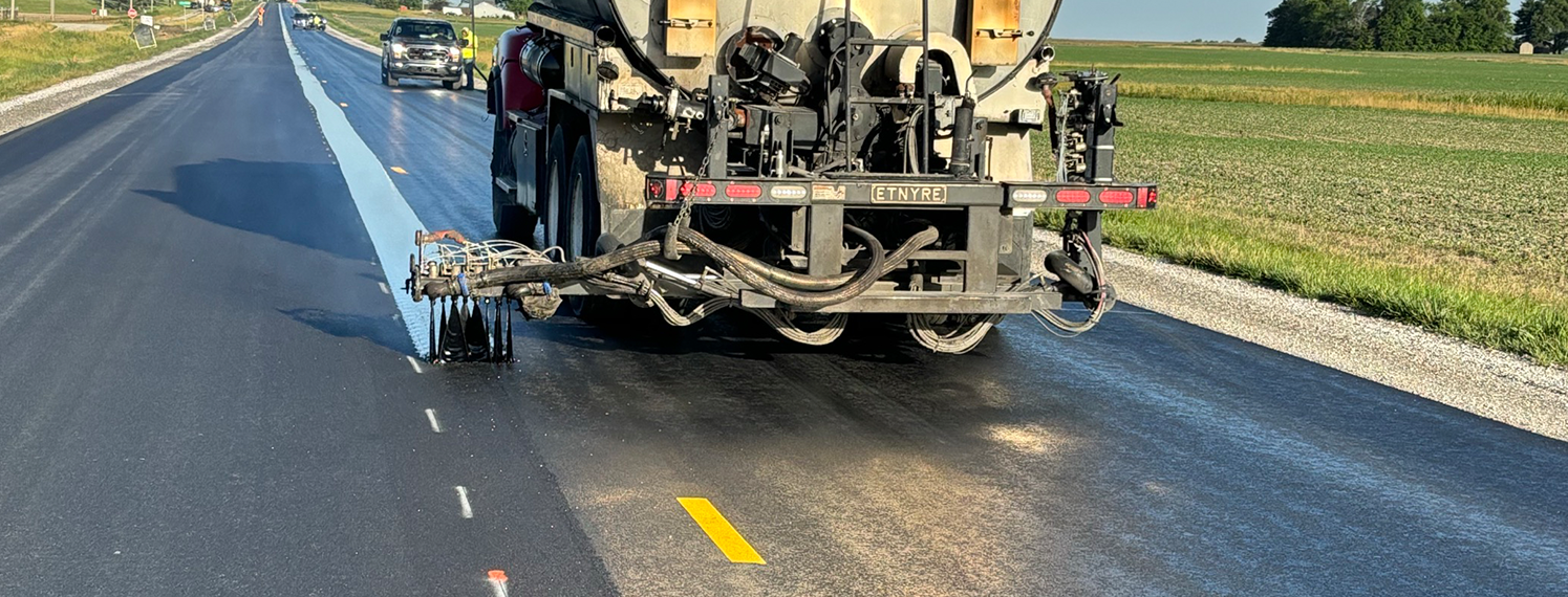 Paving truck drives down the road spraying a thing band of asphalt on the centerline