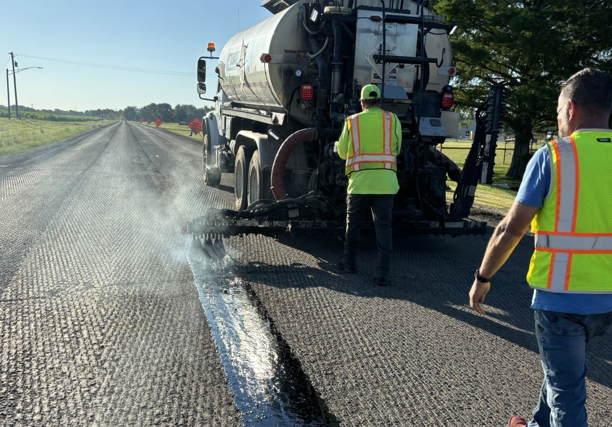 two men in construction gear walk behind a truck on a road that is being paved with J-Band